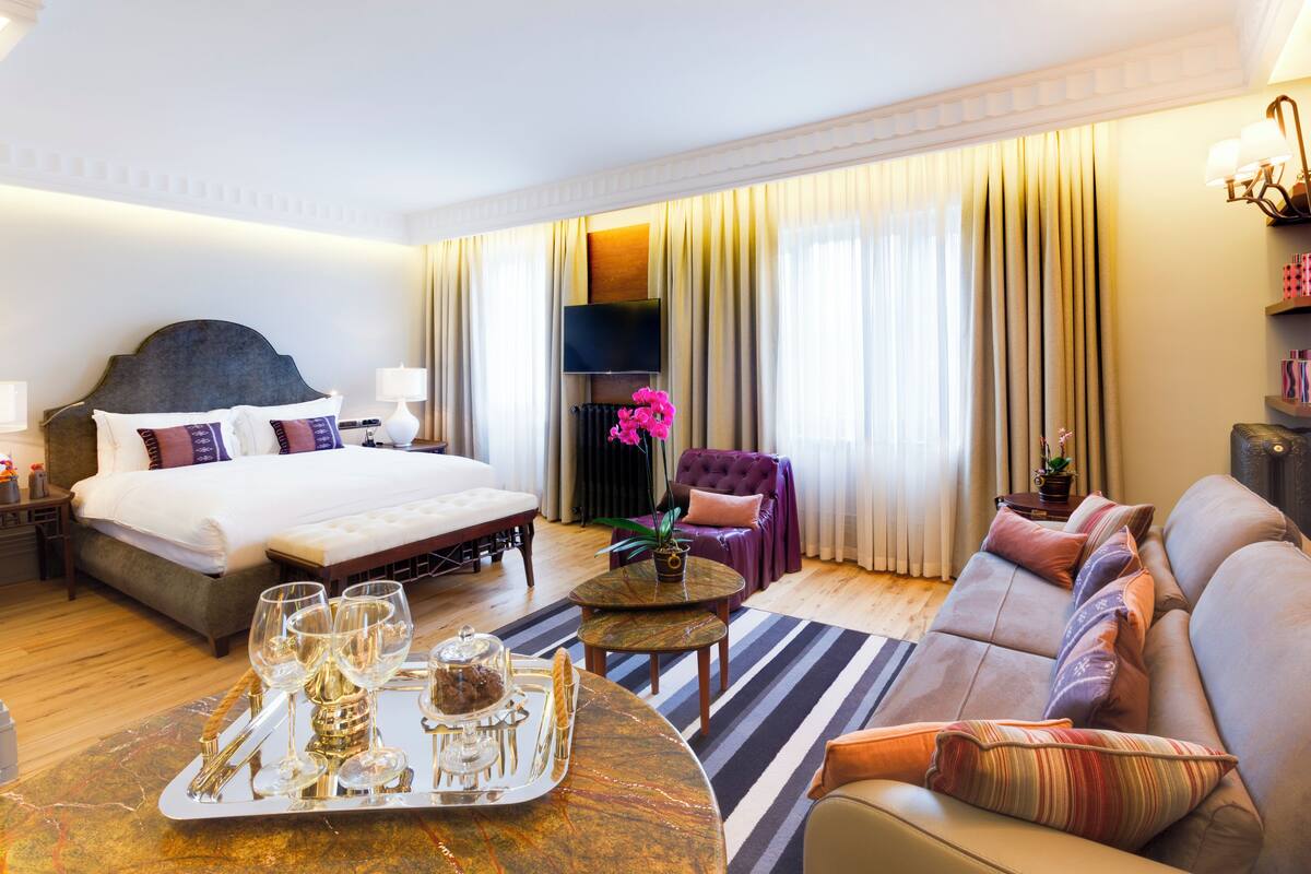 A hotel room with a bed, a sofa, a dining table with a tray of glasses and a cake, a TV, and a window with curtains.