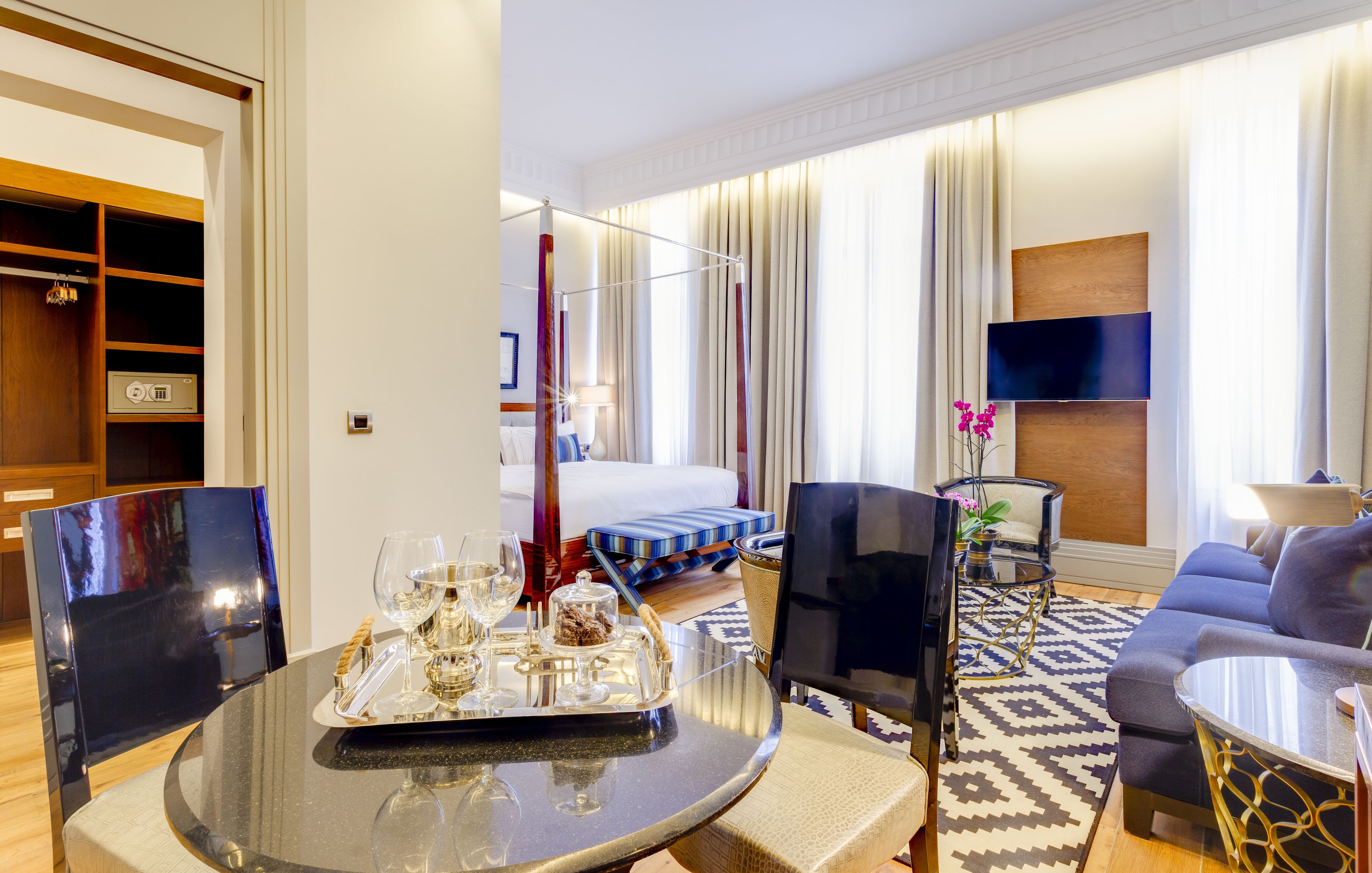 A modern hotel room with a dining area, a sofa, a TV, and a bed with curtains.