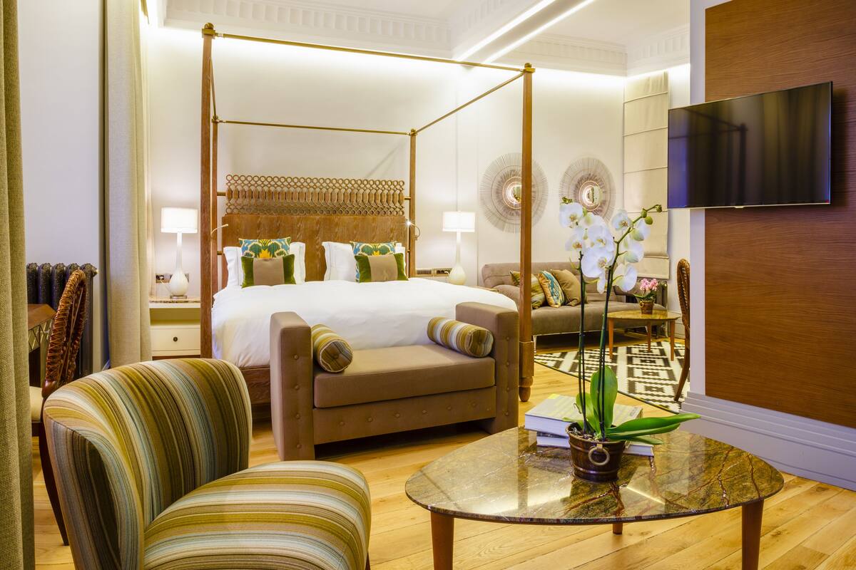 A four-poster bed with a wooden headboard, a sofa, a striped armchair, a glass coffee table with a potted orchid, and a flat-screen TV mounted on a wooden panel wall.