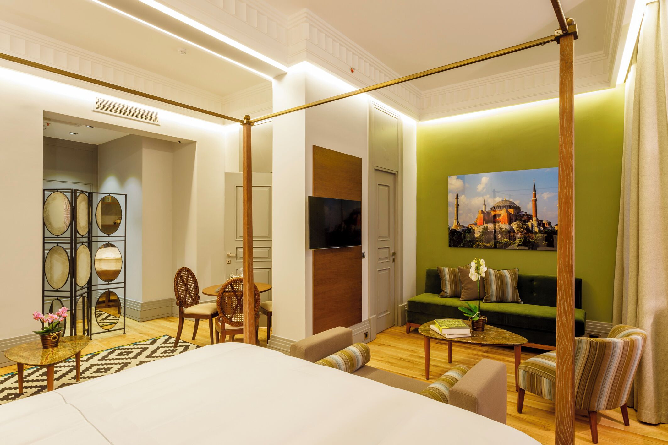 Una camera d'albergo con un letto grande, una parete verde decorativa, una televisione, un divano e un'area pranzo con sedie e tavolo.