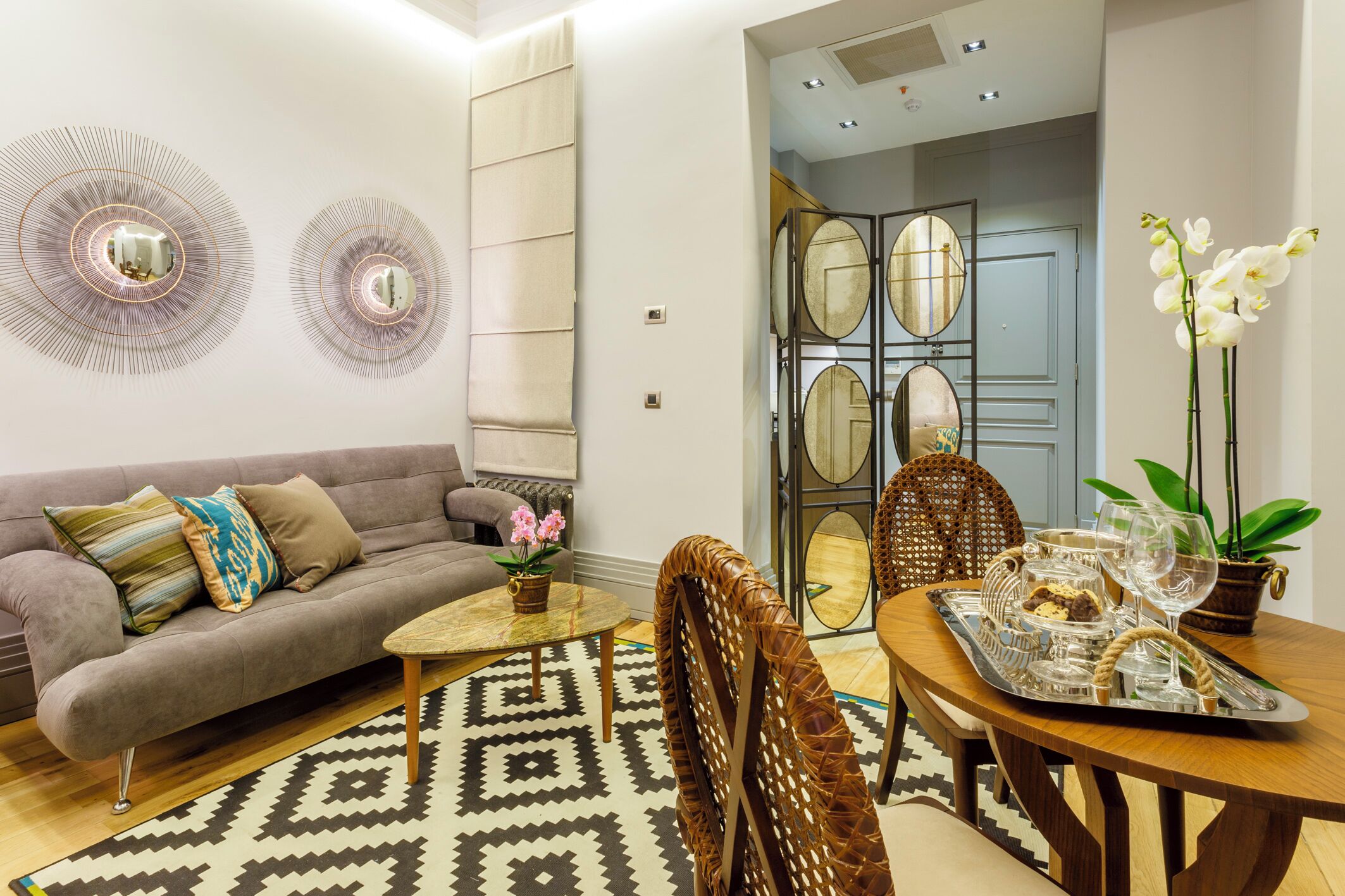 A modern living room with a sofa, coffee table, dining table, chairs, decorative mirrors, and a patterned rug.