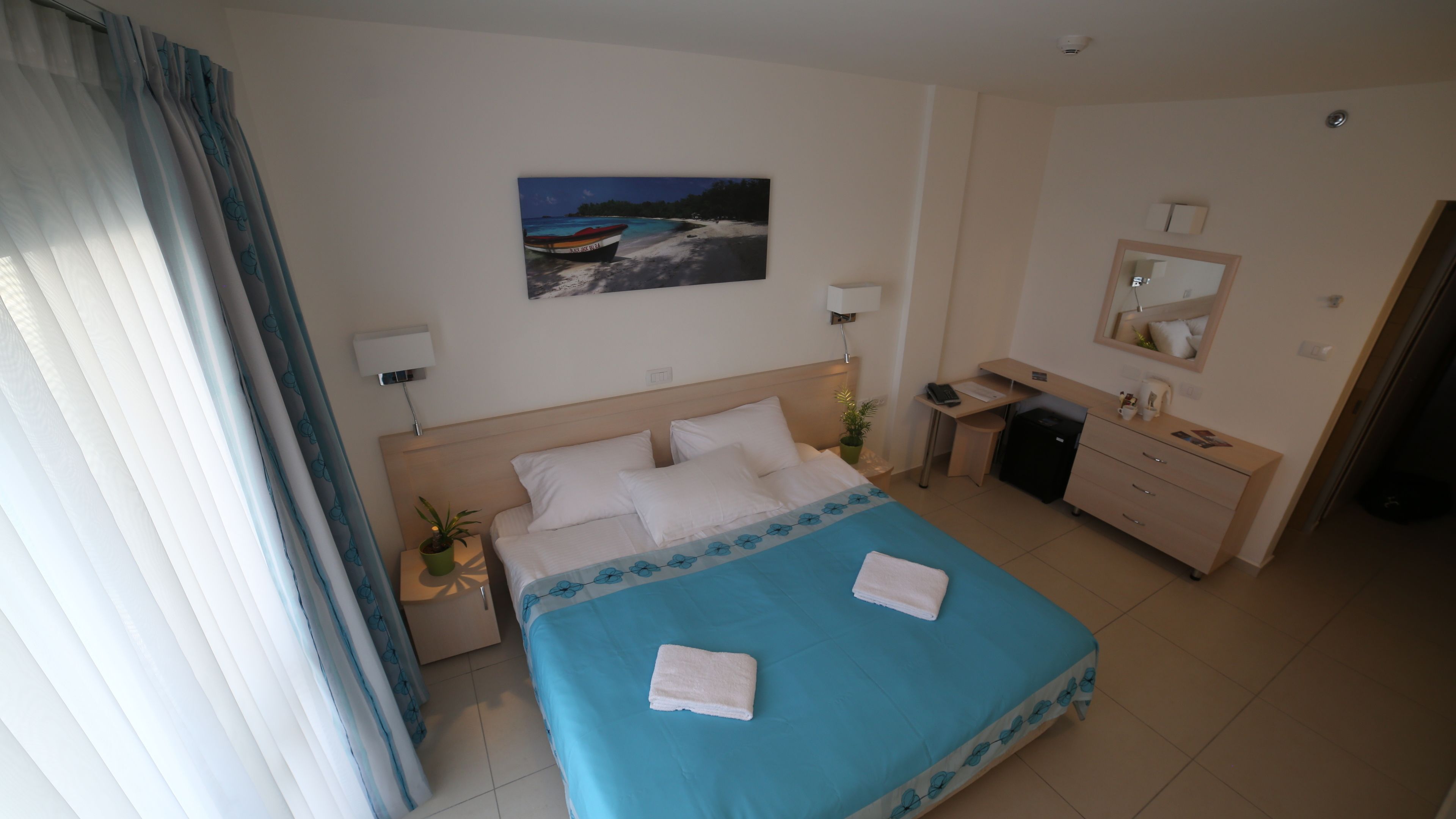 Quarto de hotel com cama, escrivaninha, cadeira, mesinha, planta em vaso e um quadro na parede.