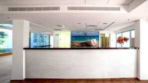 Un comptoir d'accueil au design ondulé et texturé, une affiche représentant « REGINA GOREN », et une vue sur le hall d'entrée avec un mur en verre et une plante.