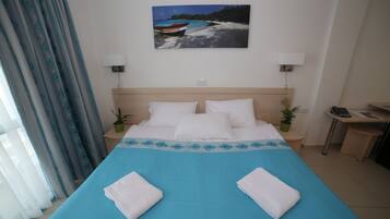 Una habitación de hotel con cama, dos mesitas de noche, escritorio, silla, lámpara, un cuadro y ventana con cortinas.