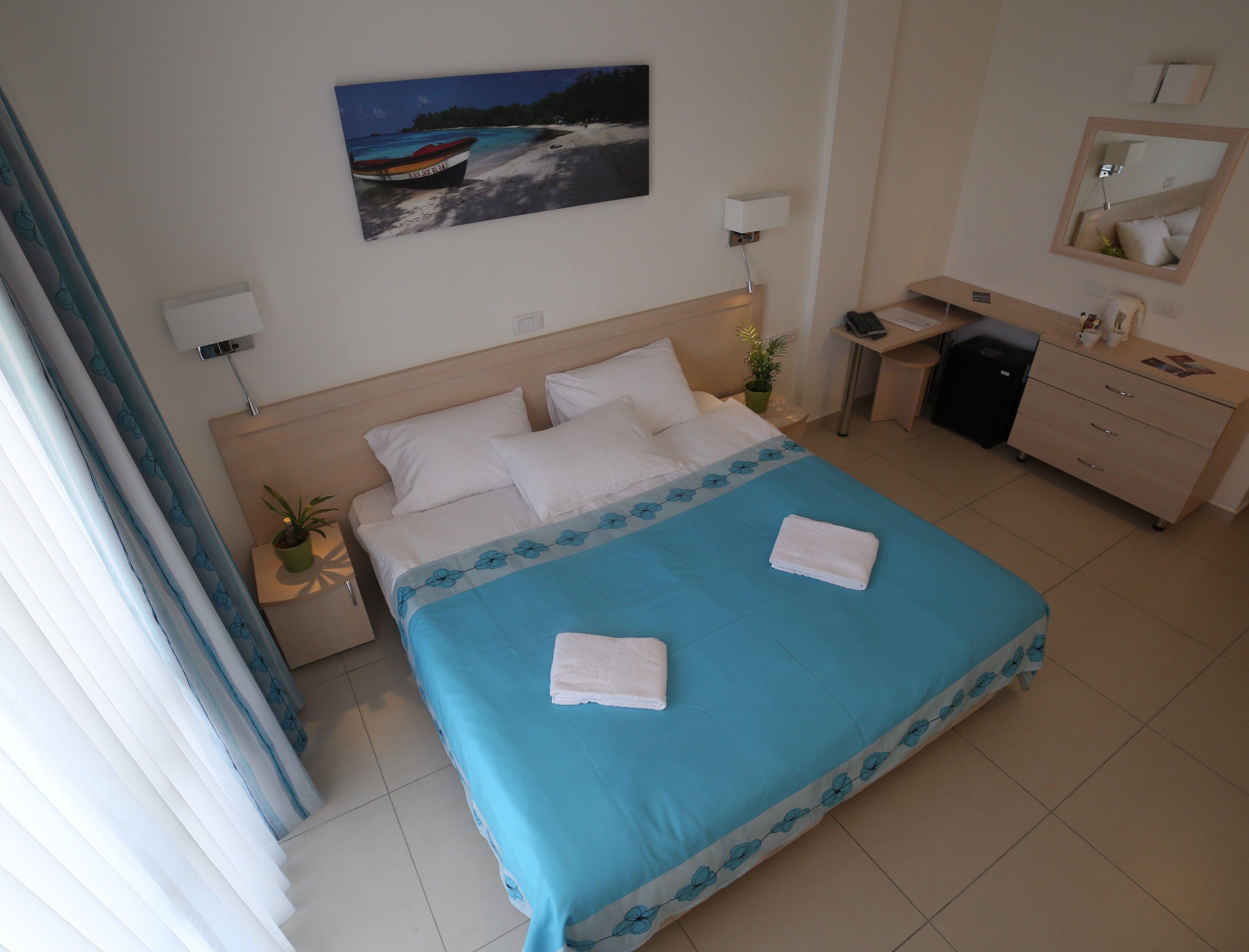 Una camera d'albergo con un letto, una scrivania, una sedia, un comò, uno specchio, una pianta in vaso e un quadro appeso al muro.