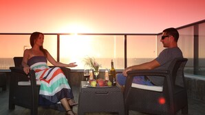 Deux personnes sont assises à une table, devant des boissons et des fruits, sur un balcon offrant une vue panoramique.