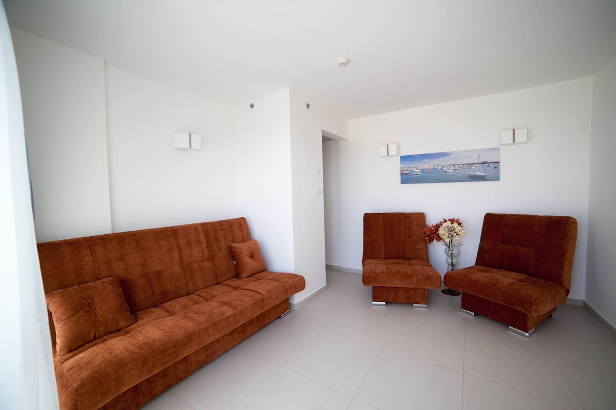 A modern living room with a large brown sofa, two armchairs, and a painting of a harbor on the wall.