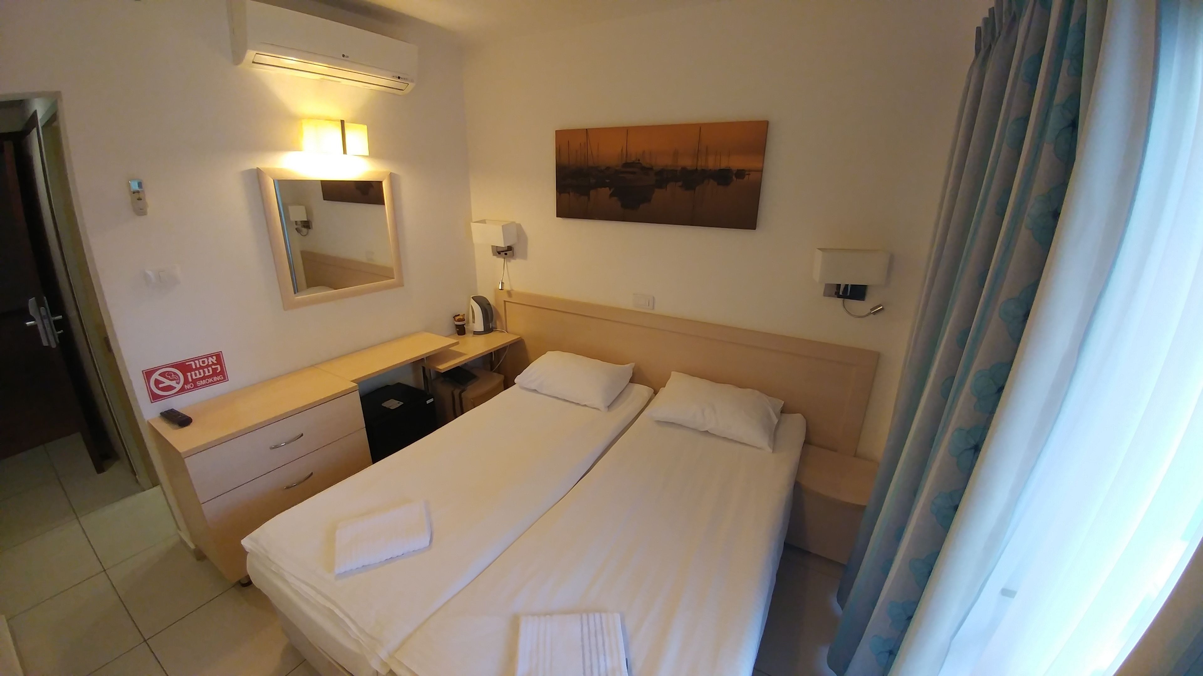 Una habitación de hotel con una cama, un escritorio con silla, un espejo, un cuadro en la pared y un cartel de no fumar.