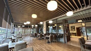 A modern restaurant with wooden flooring, large windows, and pendant lighting.