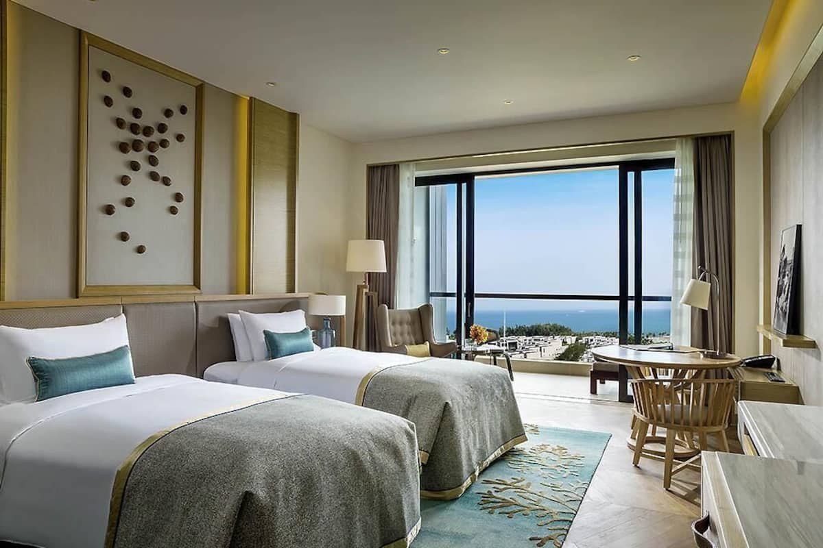 Une chambre d’hôtel avec deux lits, un bureau, une chaise, une grande fenêtre donnant sur un beau paysage et un élément décoratif mural.