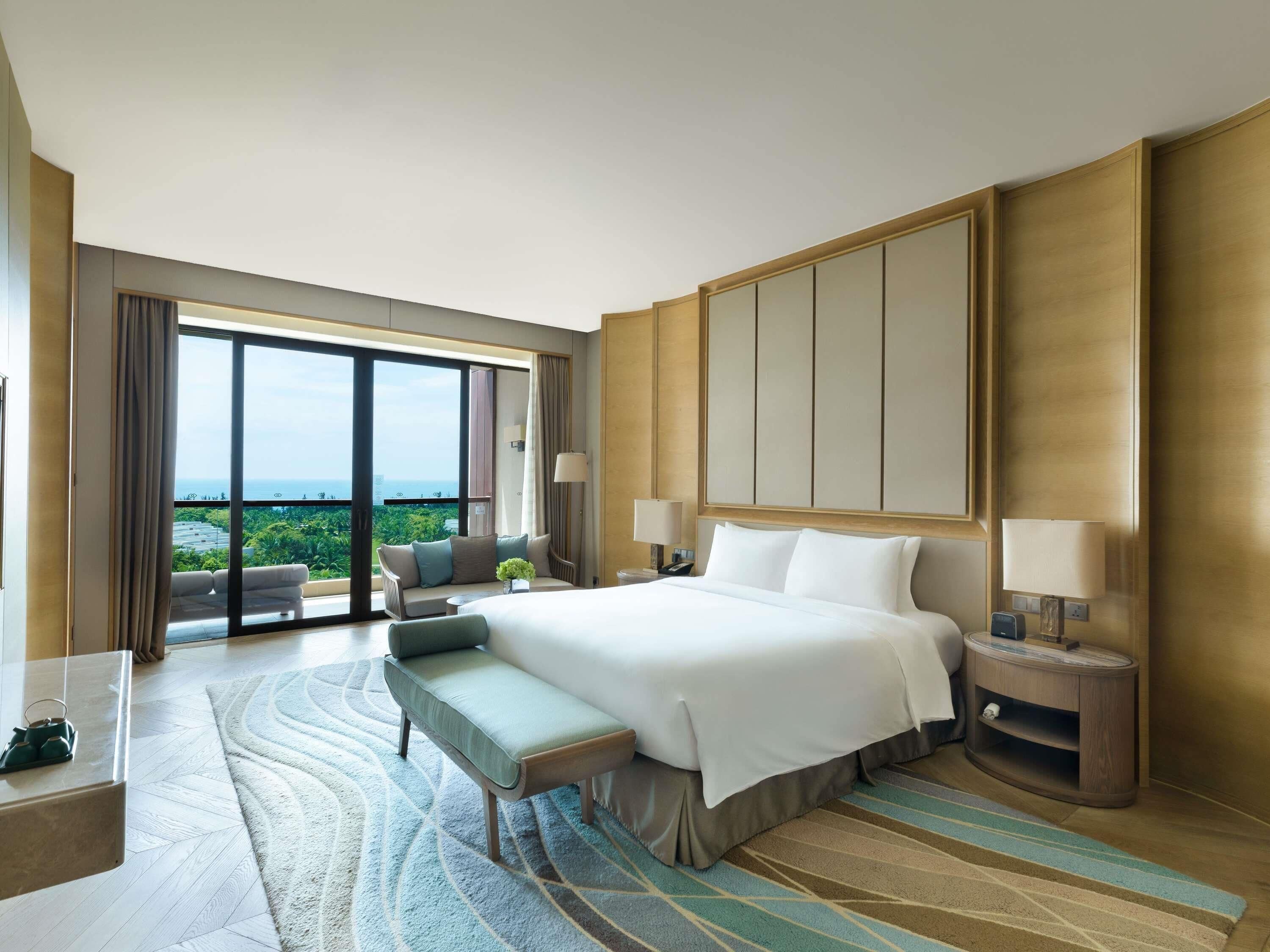 Habitación de hotel con una cama grande, un banco, una mesita de noche y vistas al exterior.