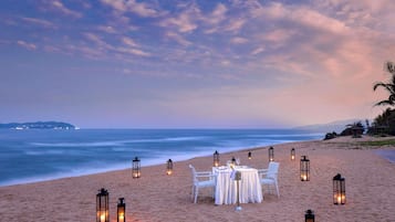 Ein Essbereich am Strand mit Tischen und Stühlen, umgeben von Laternen, unter einem Dämmerhimmel.