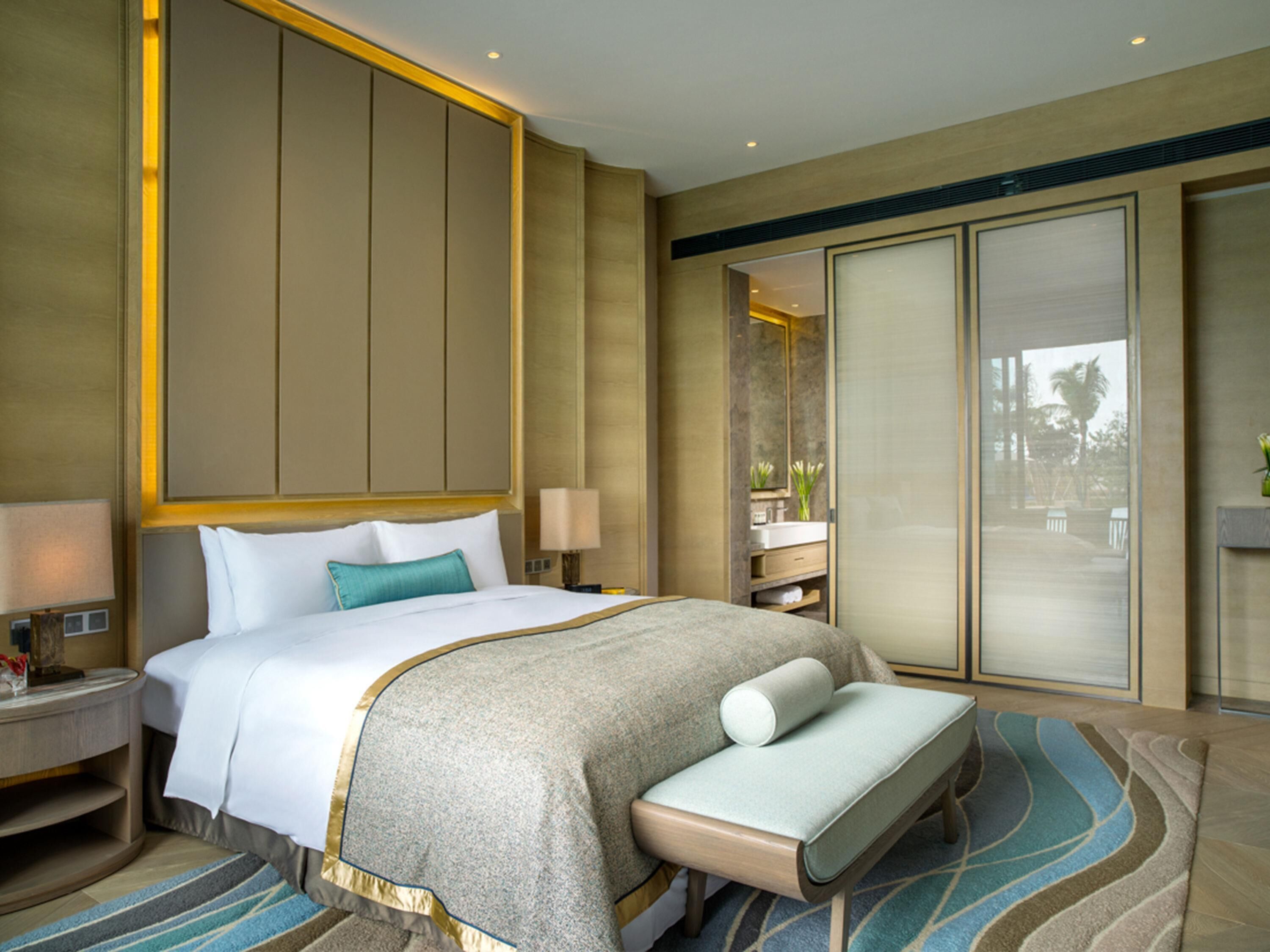 A hotel room with a large bed, a bench, a nightstand, a bedside table, a mirror, a bathroom with a sink and mirror, and a view of the outdoors.