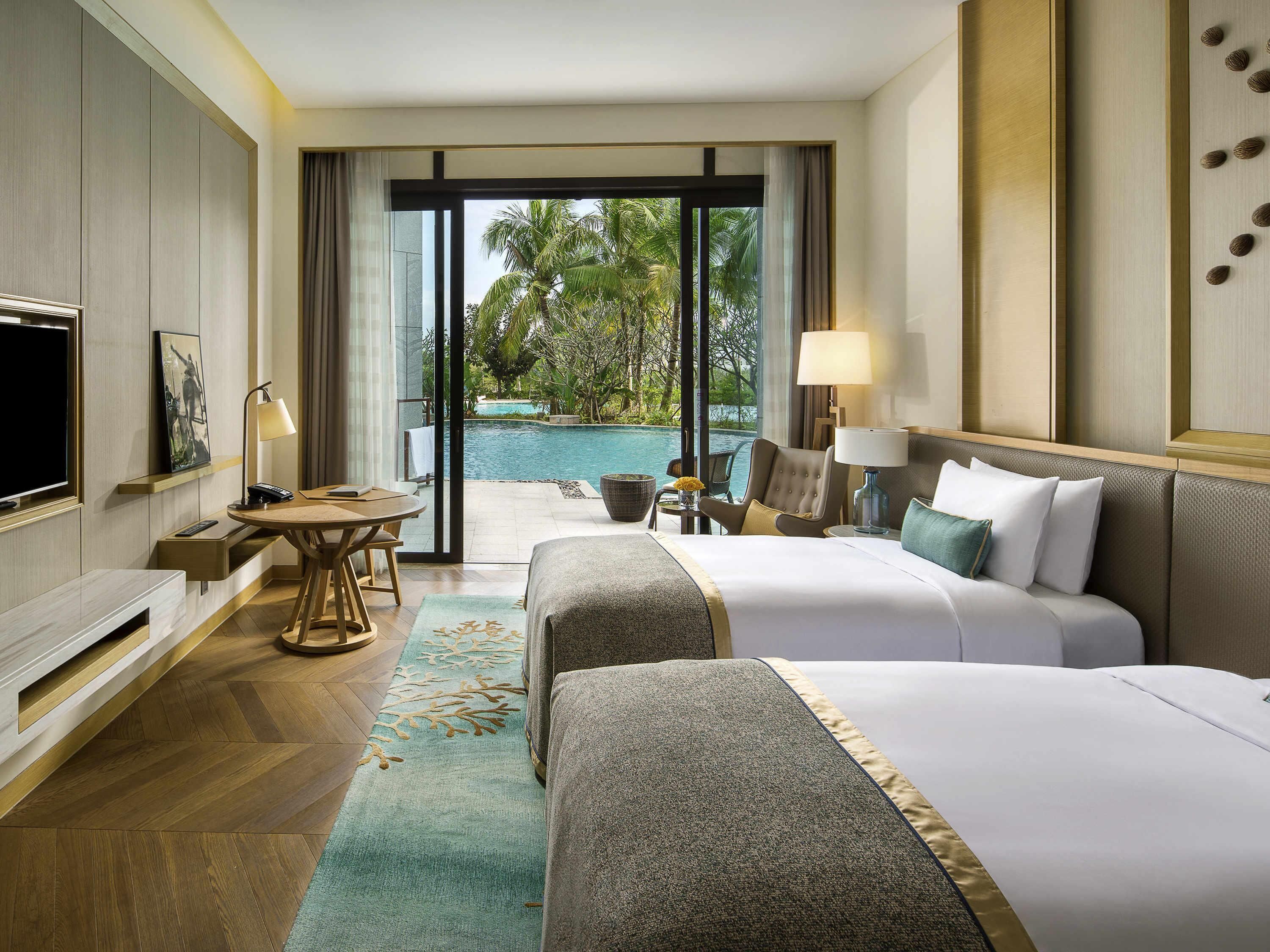 A hotel room with two beds, a TV, a desk with a chair, a lamp, and a view of a swimming pool.