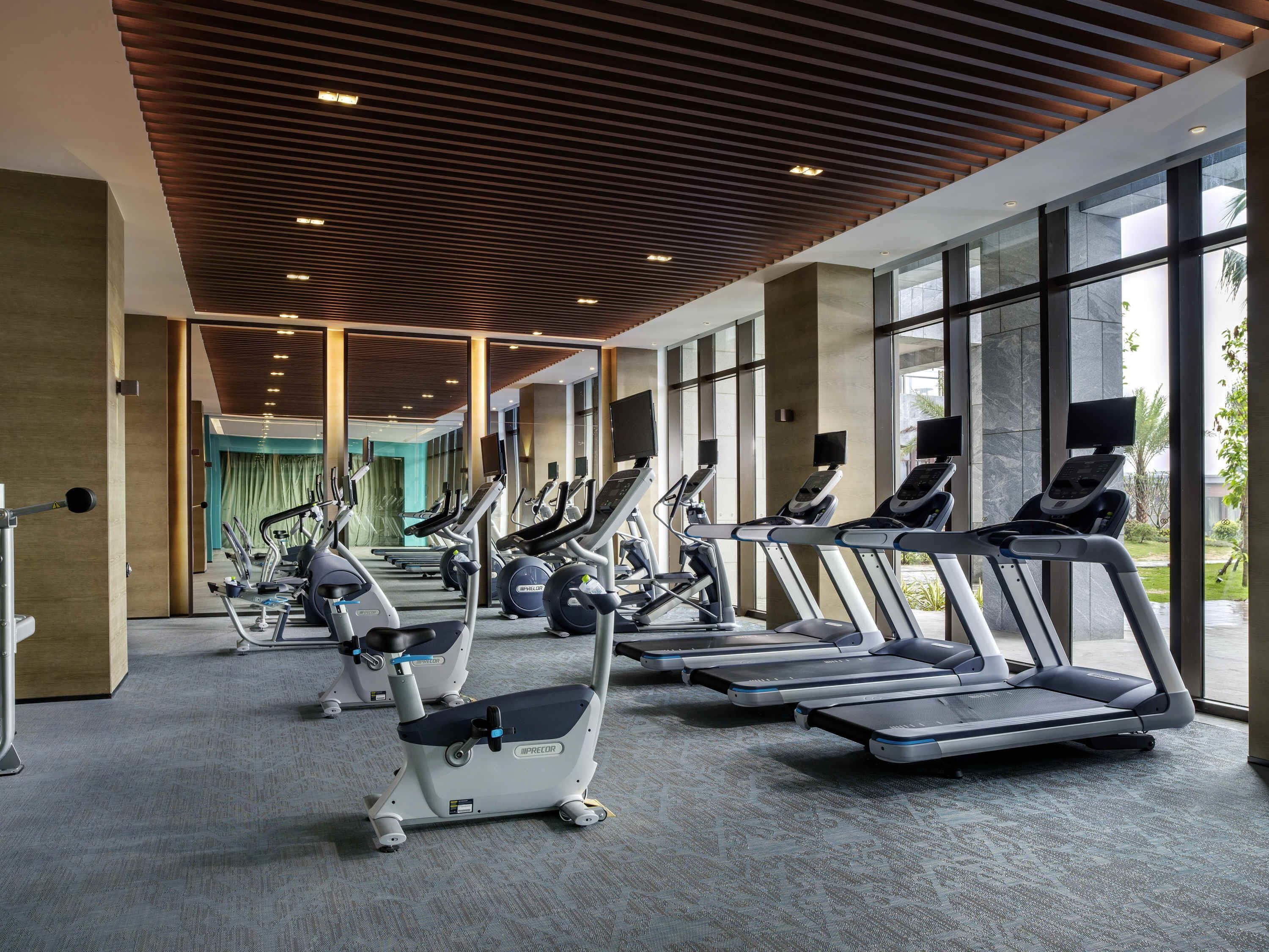 Une salle de sport équipée de plusieurs tapis de course et vélos d’exercice, dotée de grandes fenêtres et d’un design moderne au niveau du plafond.