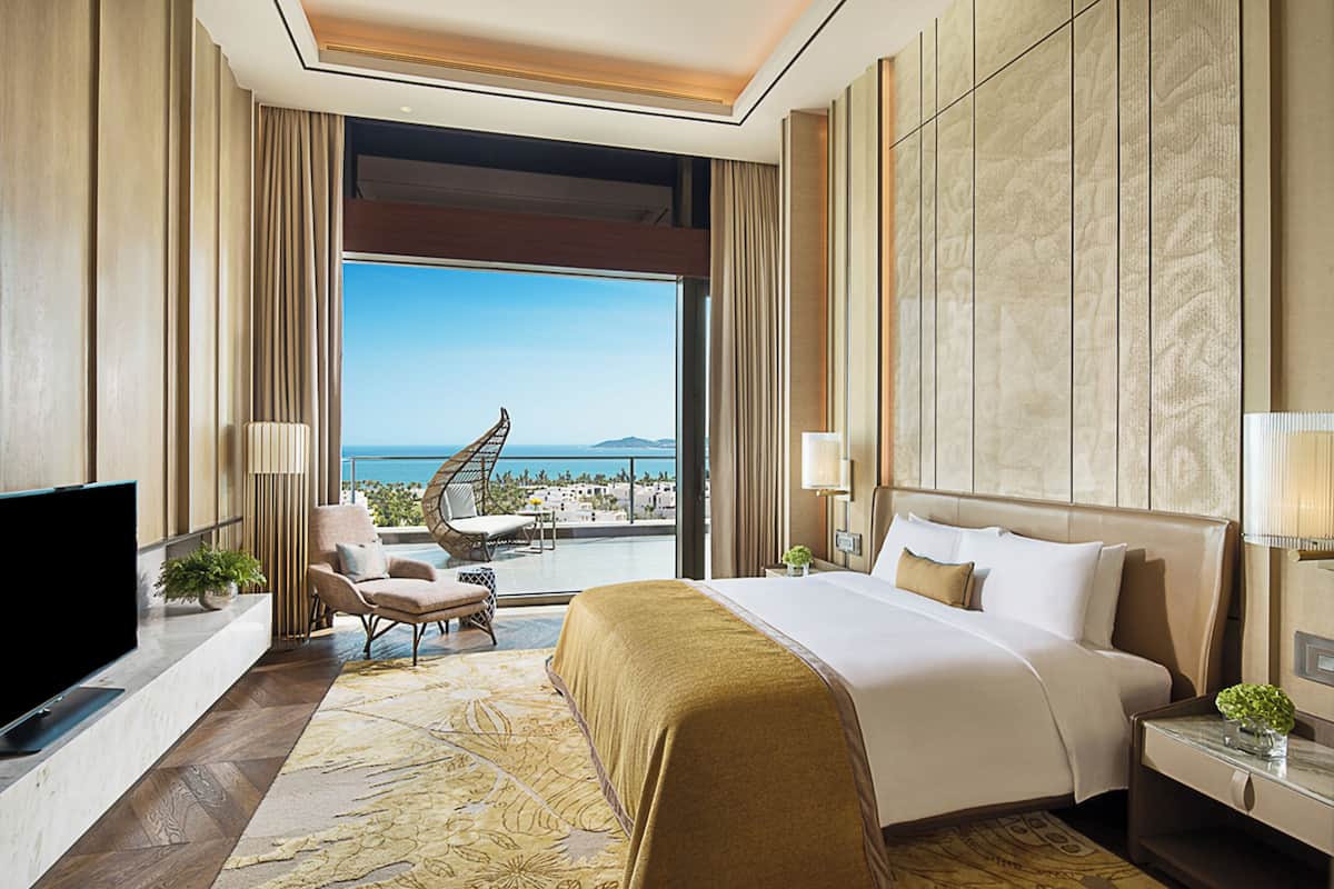 Habitación de hotel con una cama grande, televisión, balcón con vistas al mar y una zona de estar con una silla y una mesa.
