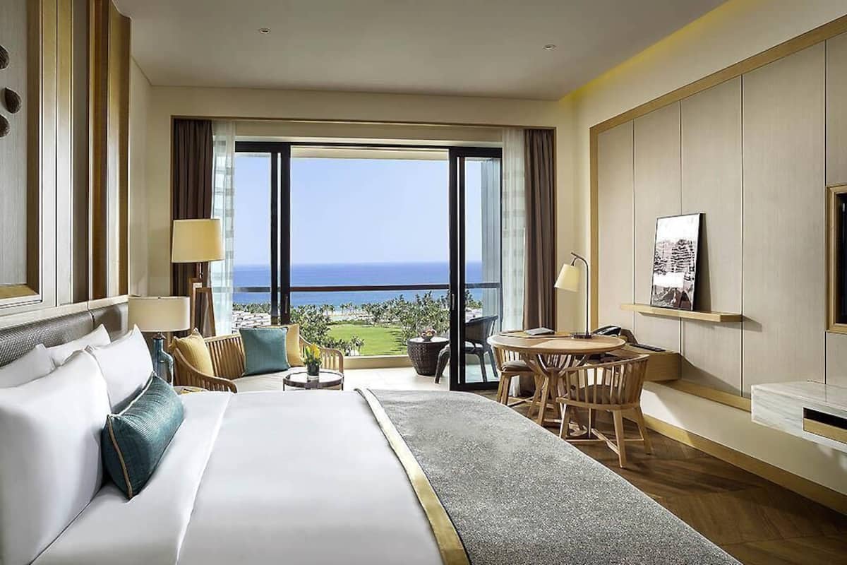 Une chambre d’hôtel avec un grand lit, un balcon avec vue, une petite table avec des chaises et un bureau avec un fauteuil.