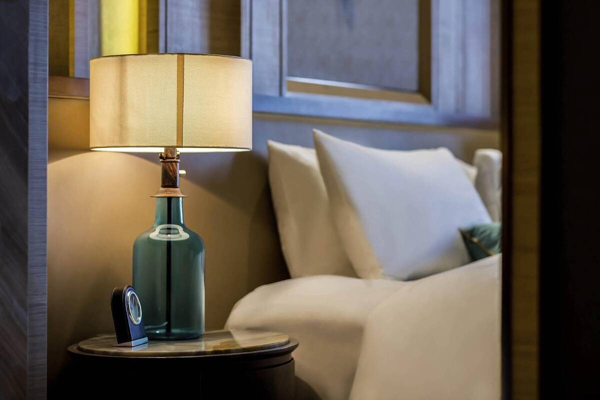 Een lamp op een nachtkastje, een bed met kussens en een muur met een deur.