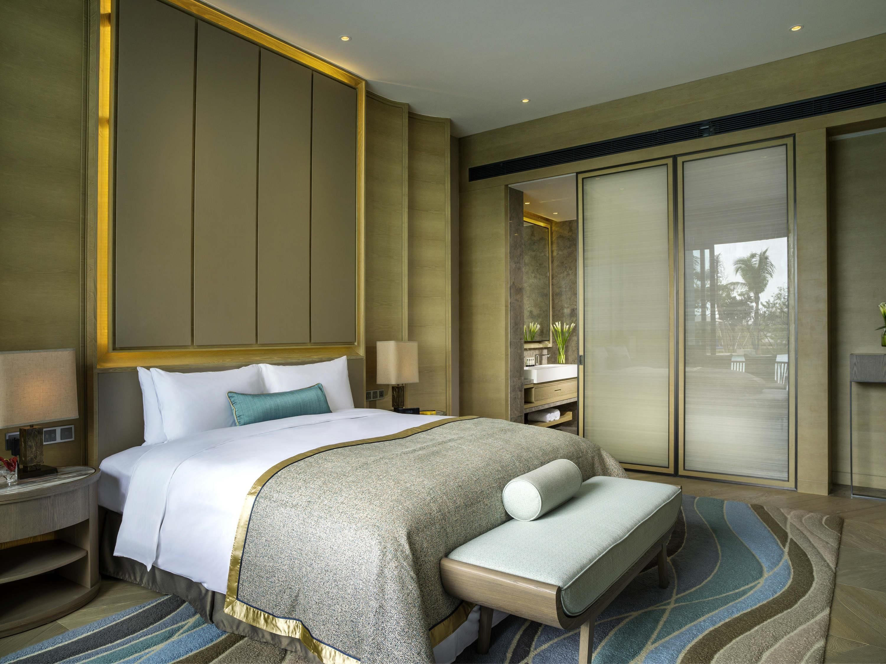 A hotel room with a large bed, bedside tables, a bench, a mirror, and a view of the outdoors.