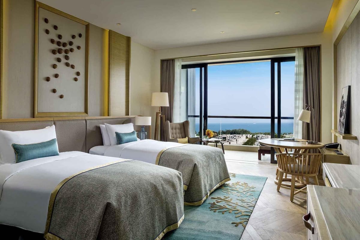 Habitación de hotel con dos camas, un escritorio, una silla, un ventanal con vistas y una obra de arte enmarcada en la pared.