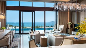 Ampla sala de estar com uma grande janela com vista para uma piscina e o mar. Há uma área de jantar com uma mesa comprida e cadeiras, e um espaço de estar com sofás e uma mesa de centro.