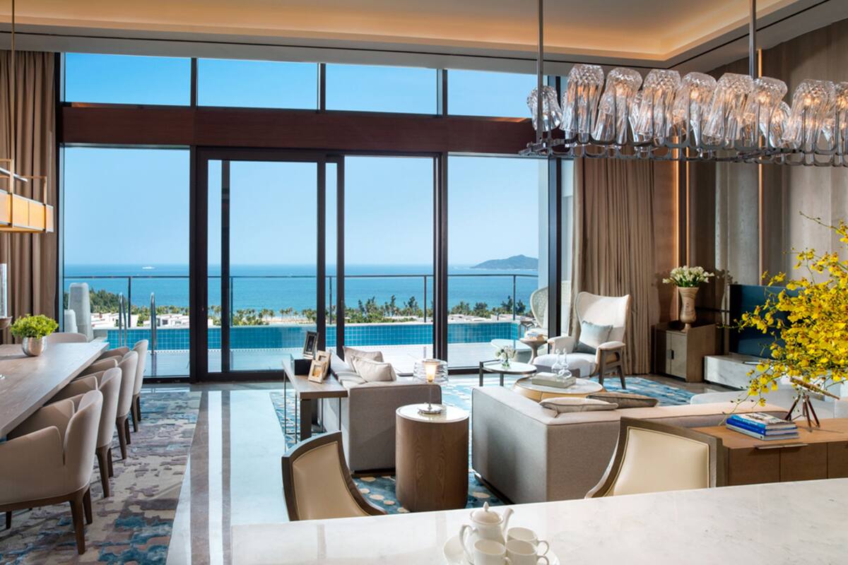 Ein geräumiger Wohnbereich mit großem Fenster mit Blick auf Pool und Meer. Es gibt einen Essbereich mit einem langen Tisch und Stühlen sowie eine Sitzecke mit Sofas und einem Couchtisch.