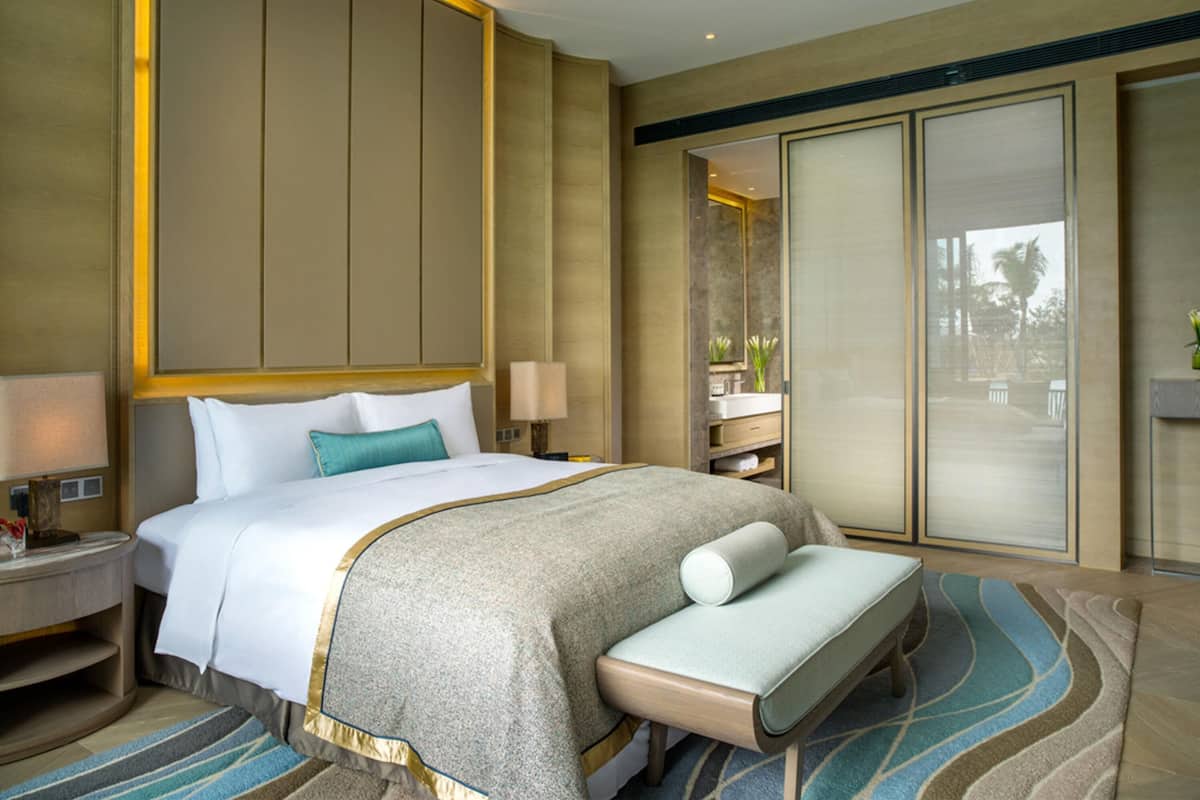 Une chambre d’hôtel avec un grand lit, une banquette, une table de chevet, une commode, un miroir et une salle de bain équipée d’un lavabo et d’un miroir, le tout offrant une vue sur l’extérieur.