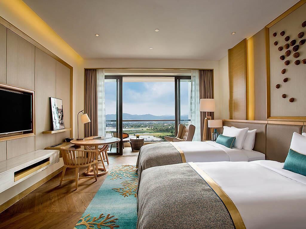 A hotel room with two beds, a TV, a desk with a chair, a view of the outdoors, and a large window.