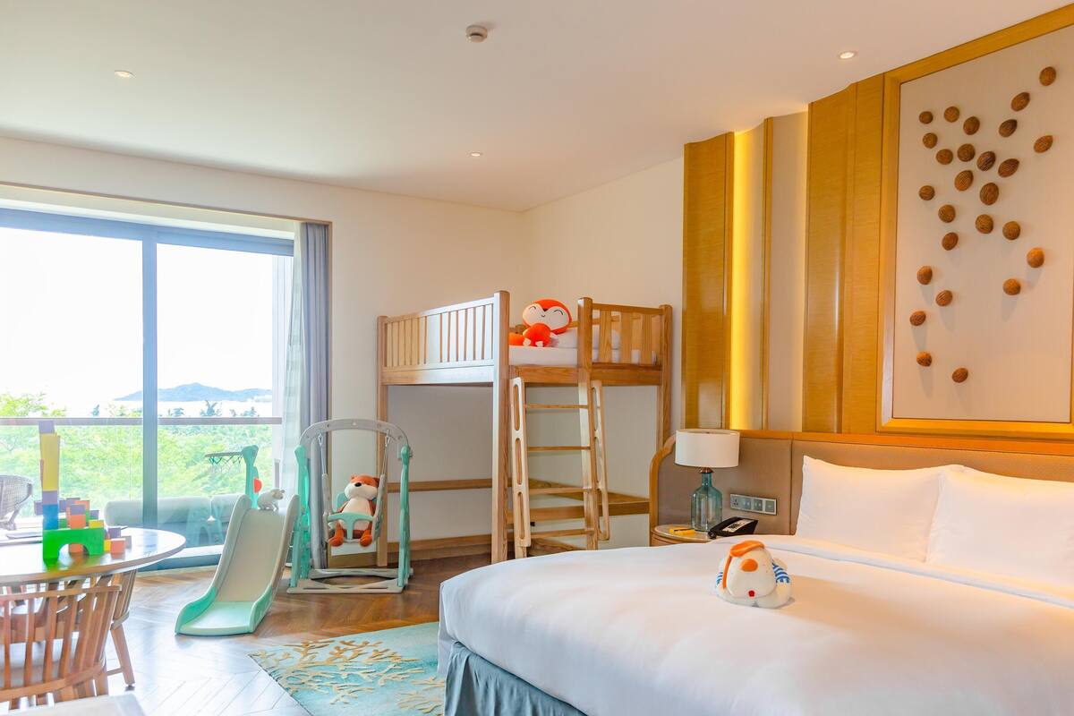 Ein Hotelzimmer mit einem Bett, einem Etagenbett, einem Schreibtisch mit Stuhl, einer Spielecke mit Spielzeug und einem großen Fenster mit Aussicht.
