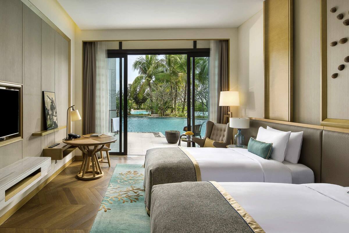 Ein Hotelzimmer mit zwei Betten, einem Fernseher, einem Schreibtisch mit Lampe und Blick auf einen Swimmingpool.