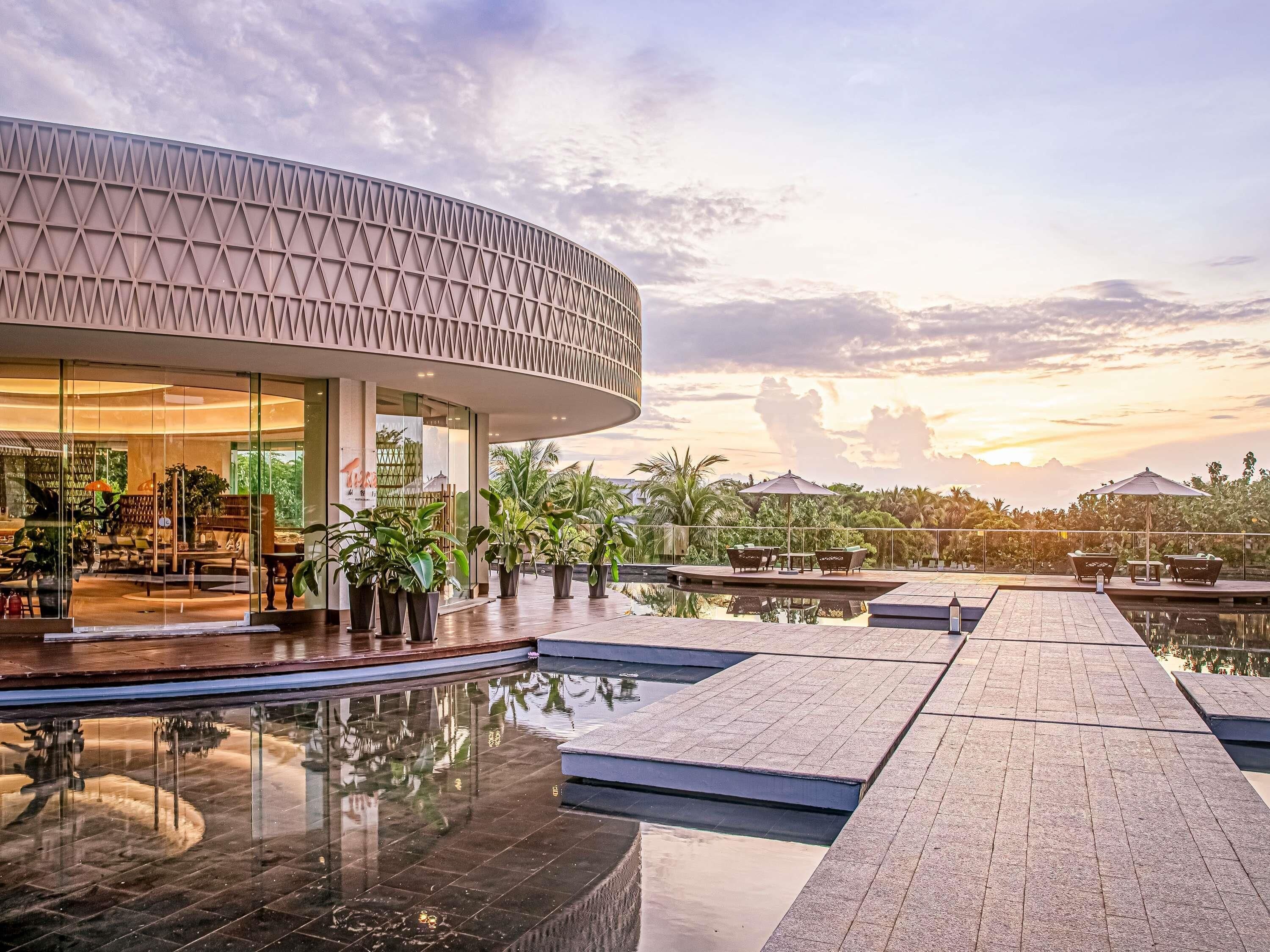 Un bâtiment moderne à la façade incurvée, doté d’un espace piscine protégé par une paroi en verre et d’un espace de détente extérieur.