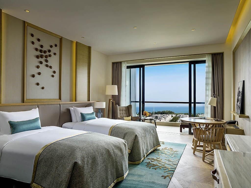 A hotel room with two beds, a desk, a chair, a large window with a view, and a decorative wall piece.