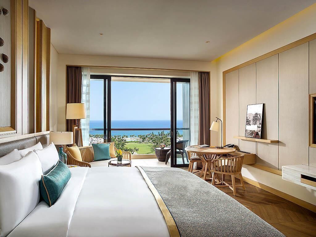 Quarto de hotel com cama grande, escrivaninha, cadeira e vista para o oceano.
