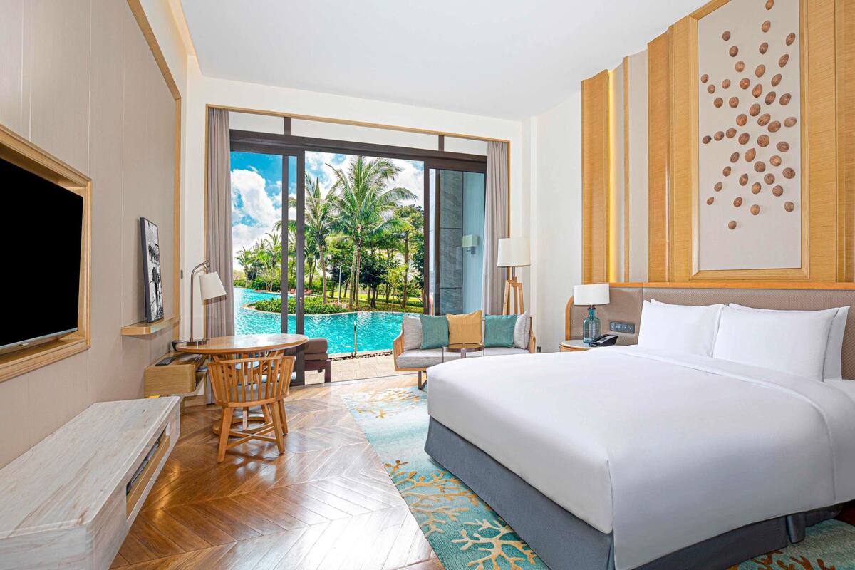 Ein Hotelzimmer mit einem großen Bett, einem Schreibtisch mit Stuhl, einem Fernseher und Blick auf einen Pool.