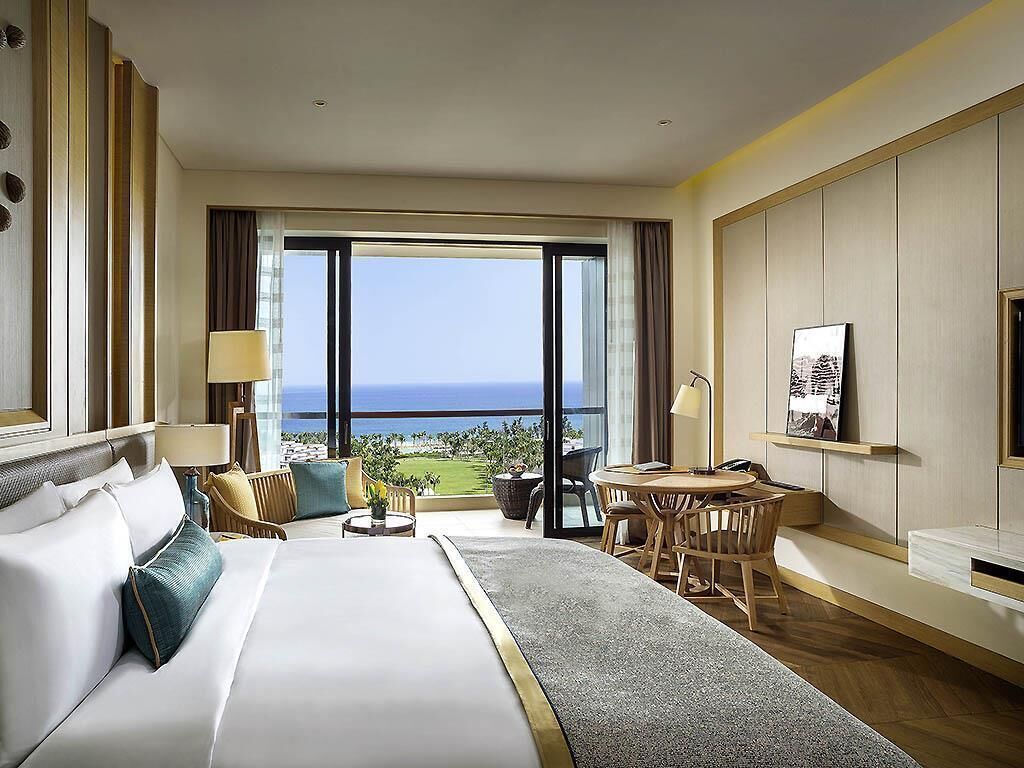 A hotel room with a large bed, a balcony with a view, a small table with chairs, and a desk with a chair.
