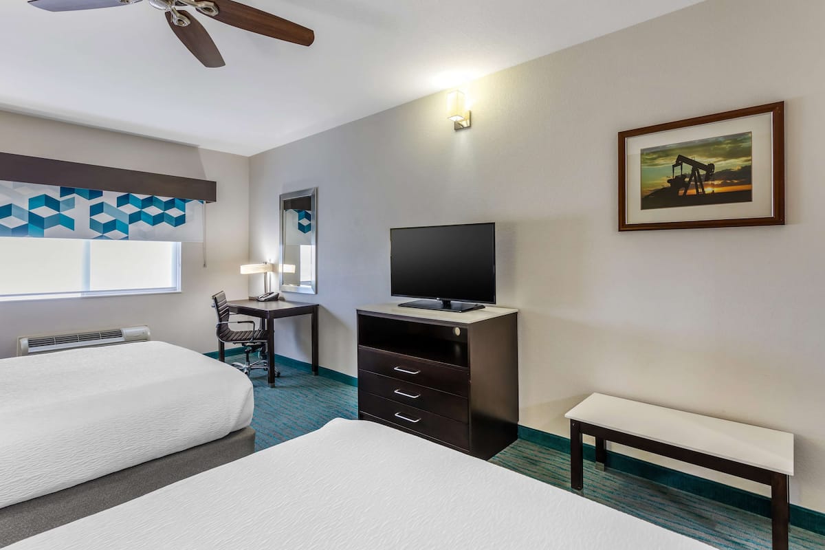 A hotel room with two beds, a desk with a chair, a television, a bench, and a painting on the wall.