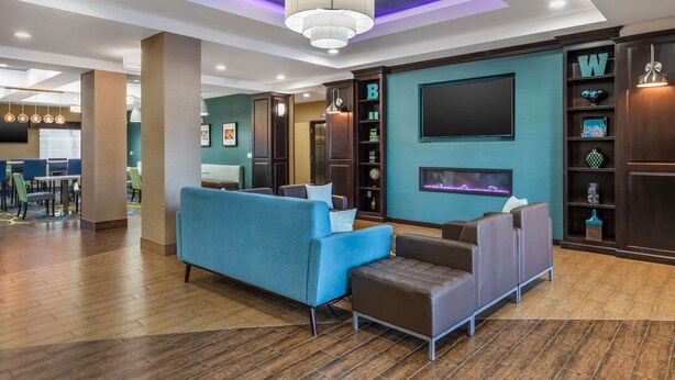 A modern lobby with a blue sofa, a brown ottoman, and a chandelier. There is a wall-mounted TV and a shelf with decorative items.