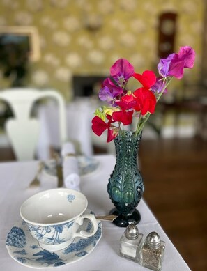Una mesa con un florero decorativo que contiene flores coloridas, una taza blanca con plato, y saleros y pimenteros.
