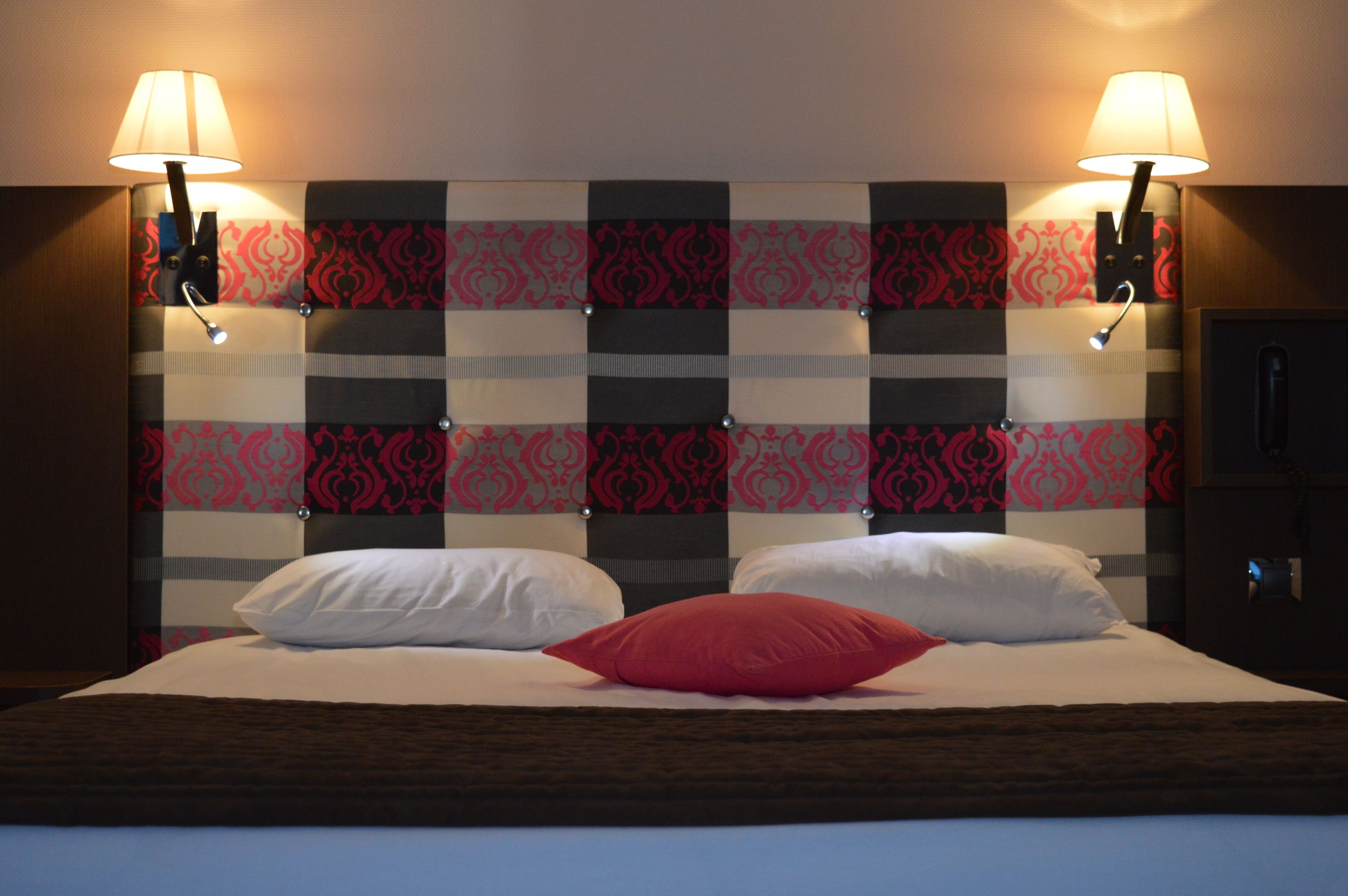 A hotel room with a checkered headboard, two bedside lamps, and a bed with white and red pillows.