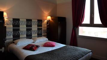 A hotel room with a bed, two pillows, a quilted blanket, a patterned headboard, a window with curtains, and wall-mounted lamps.