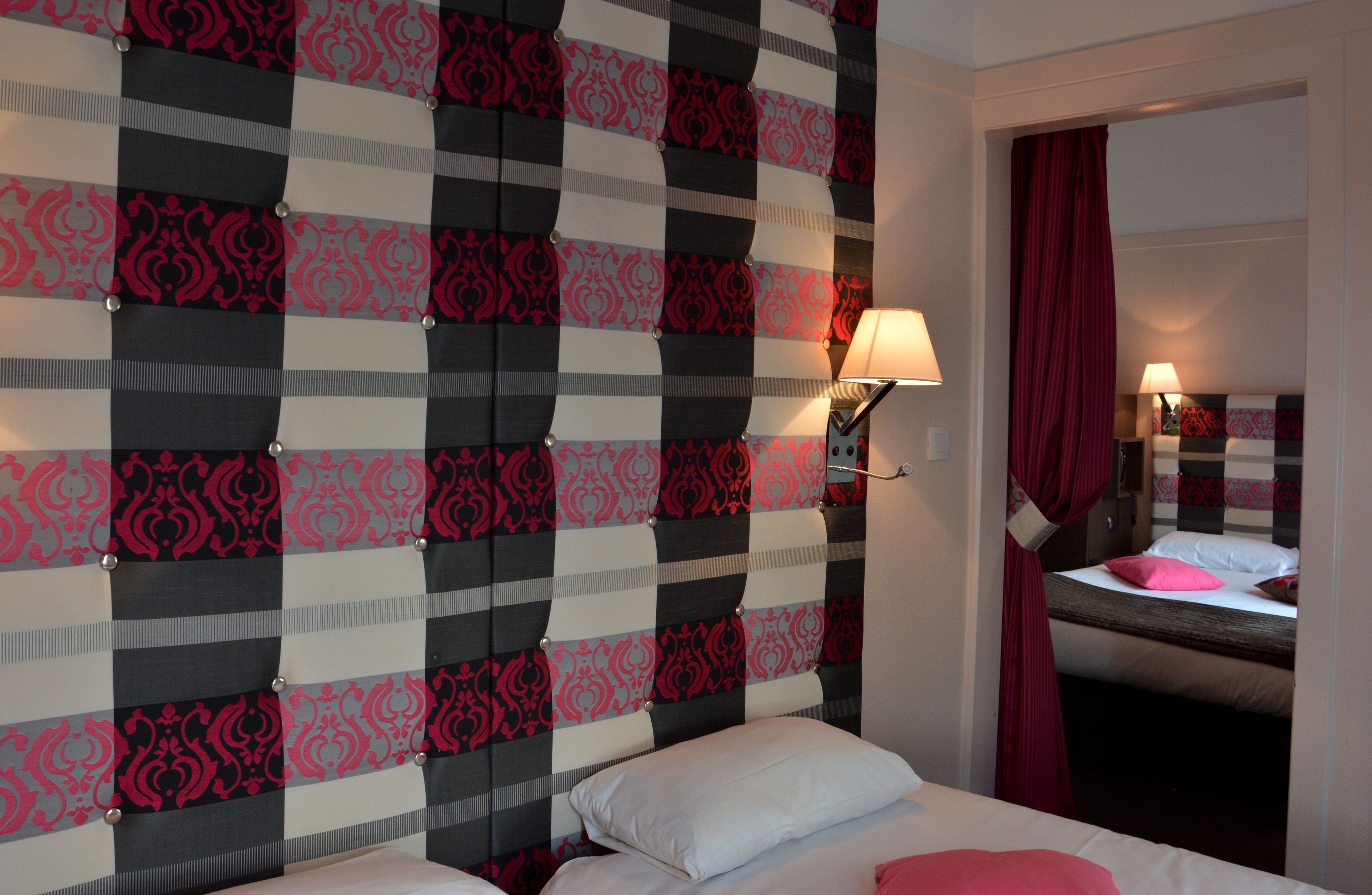 Habitación de hotel con cabecera a cuadros, cama con ropa de cama blanca y rosa, mesita de noche con lámpara y ventana con cortina roja.