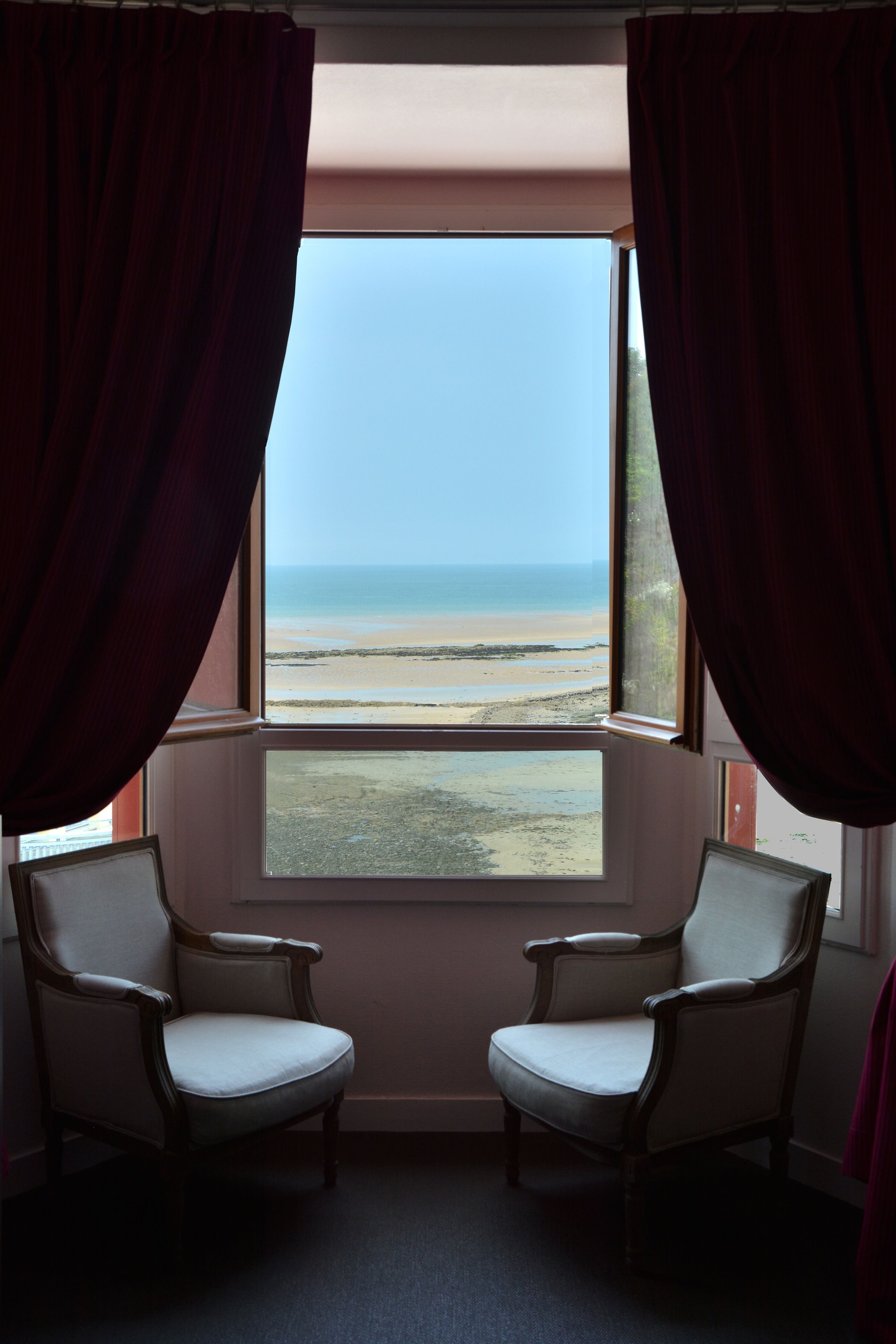 Een kamer met een groot raam dat uitkijkt op een strand, twee stoelen en uitzicht op de zee.