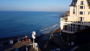 A coastal building with a balcony overlooking the sea, a parking area with cars, and a clear sky.
