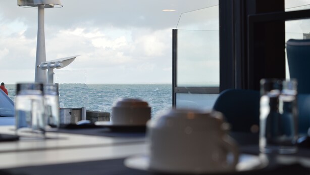A table with a view of the sea through a window, a ship's mast, and a glass of water.