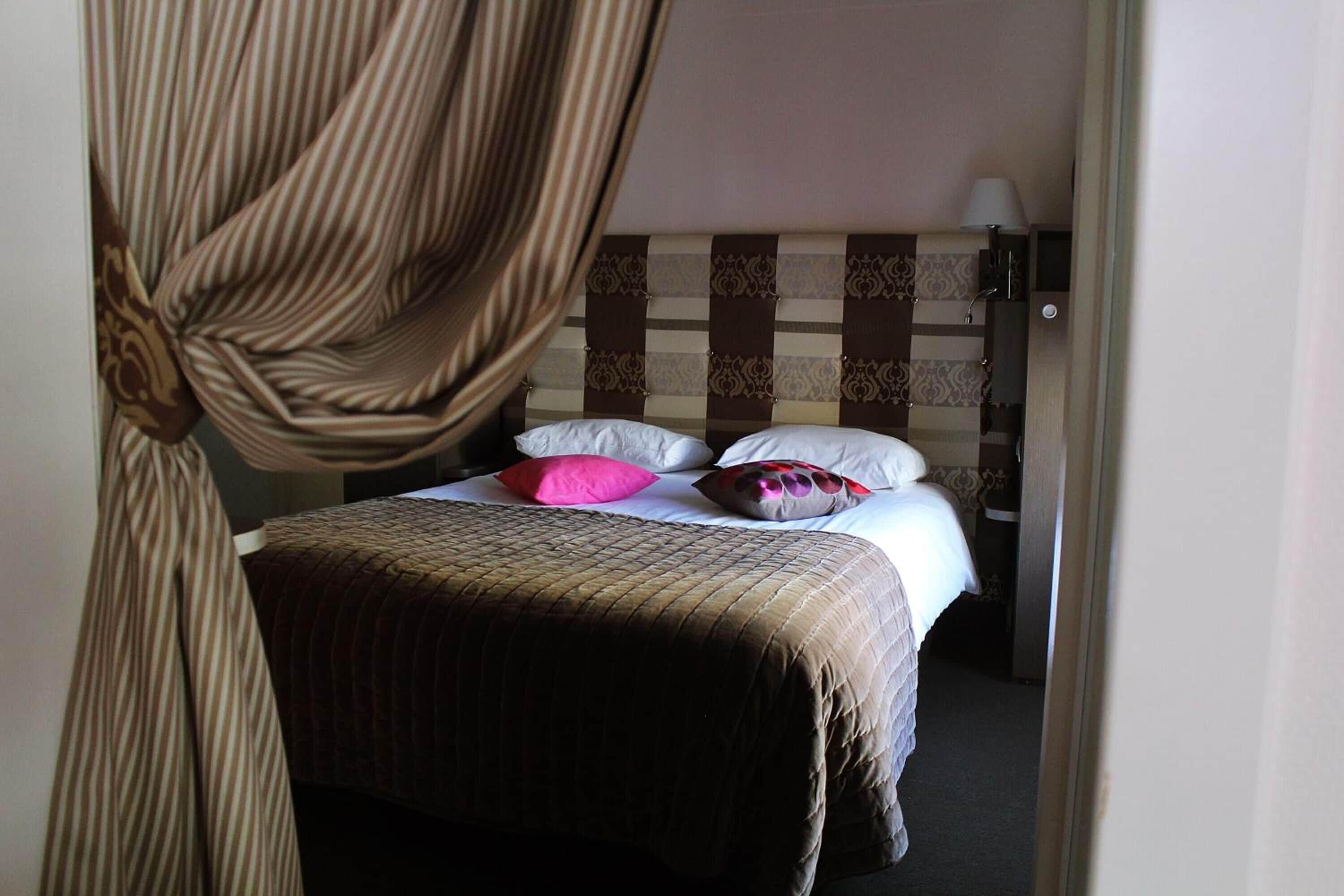 A hotel room with a bed, a headboard with a patterned design, and a curtain partially drawn.