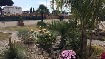 Un jardín con diversas plantas y flores, una palmera y un edificio al fondo.