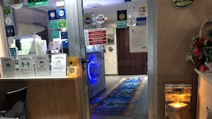A reception area with a glass door, a reception desk, and a digital screen displaying information. There are signs for Wi-Fi, Google, and American Express. A blue-lit pathway leads inside, and a small fire feature is visible on the right. - Il Giardino di Adriana (Terracina)