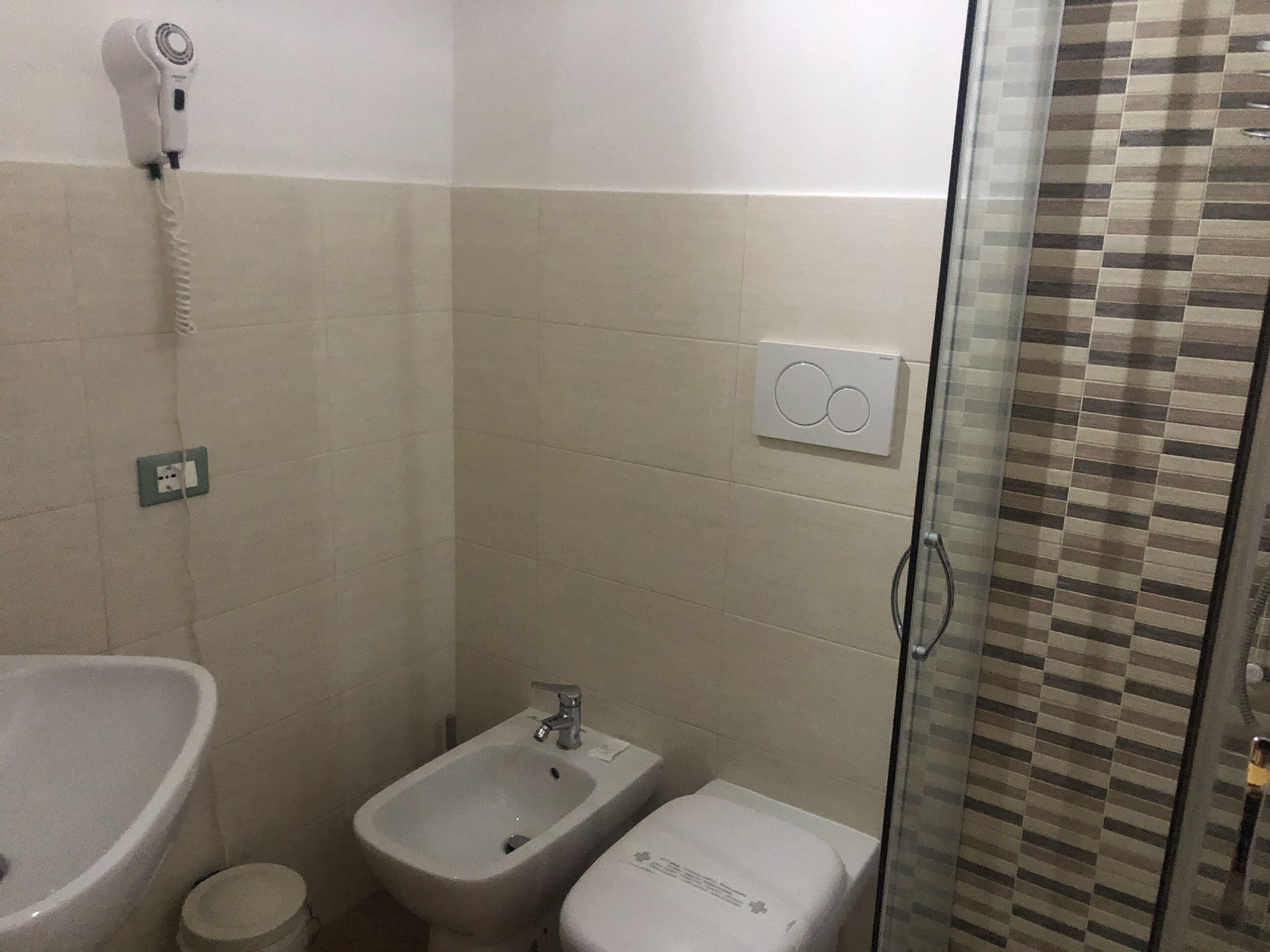 Une salle de bain équipée d’un bidet, d’un lavabo, d’un sèche-cheveux fixé au mur et d’une douche avec une porte en verre.