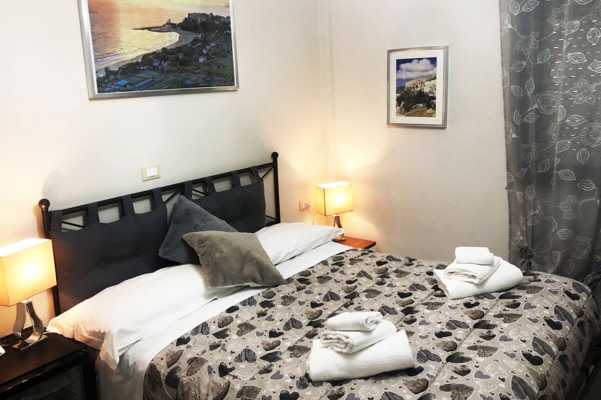 Ett hotellrum med en säng, två sänglampor, ett nattduksbord, en inramad tavla ovanför sängen och en gardin med ett blommönster.