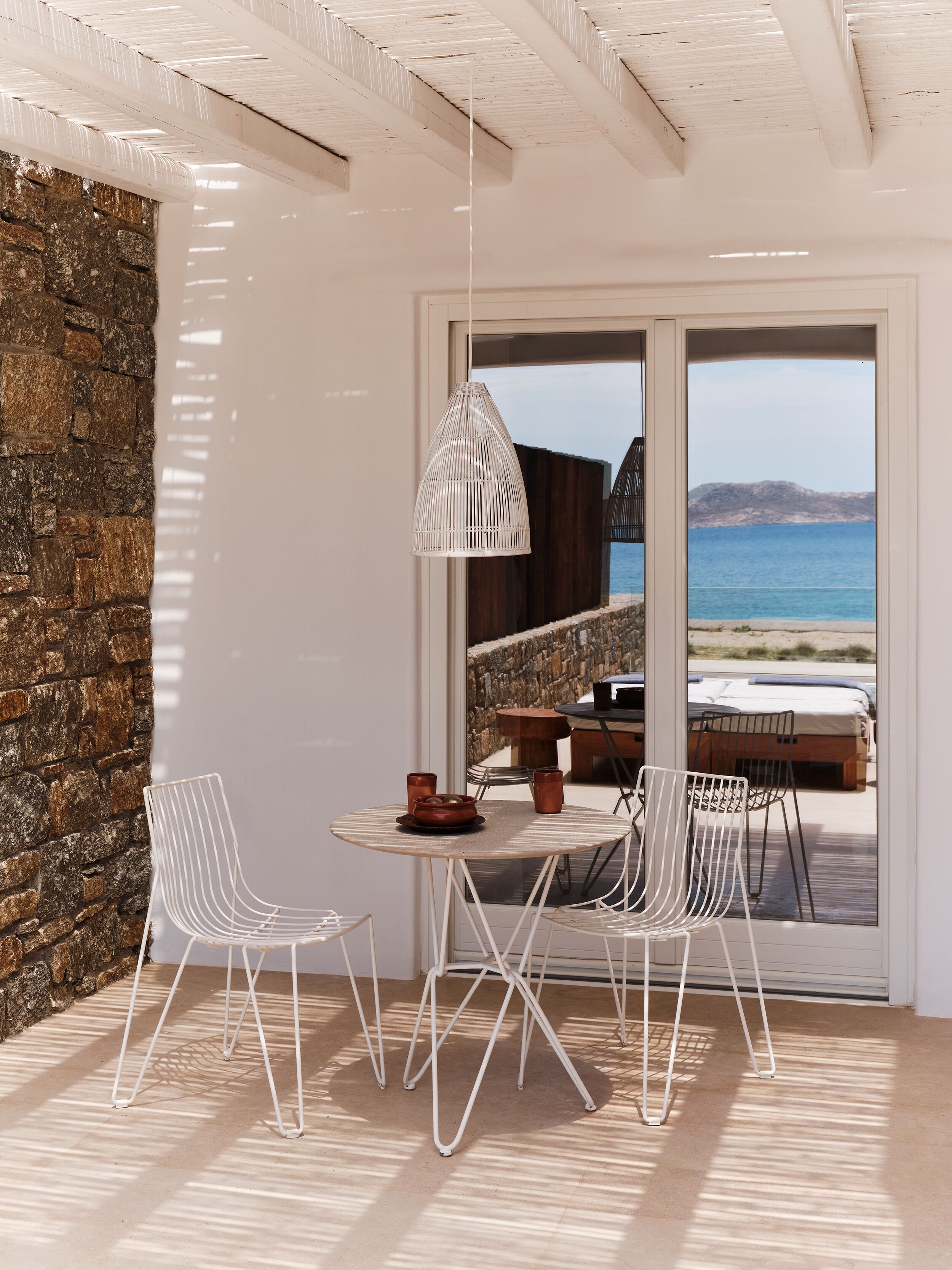 Une pièce moderne avec un mur en pierre, une table ronde entourée de chaises, un luminaire suspendu et une vue sur la mer grâce à de grandes portes coulissantes.